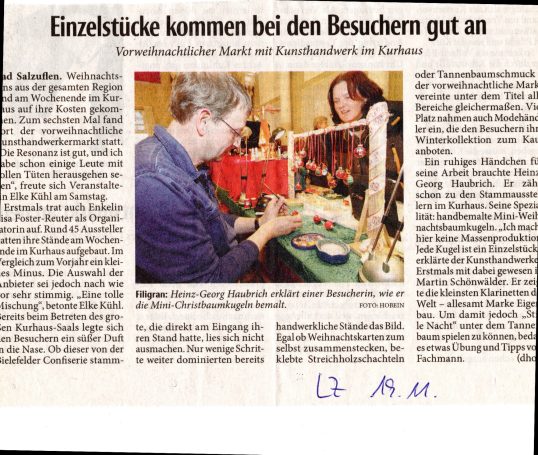 Zeitungsbericht über Kunsthandwerkermarkt. in Bad Salzuflen. Ein Mann sitzt an einem Tisch und bemalt kleine Weihnachtskugel. Eine Kundin sc